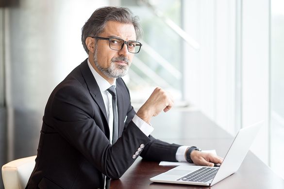 hombre de pelo canoso con gafas y trajeado en oficina con ordenador portátil